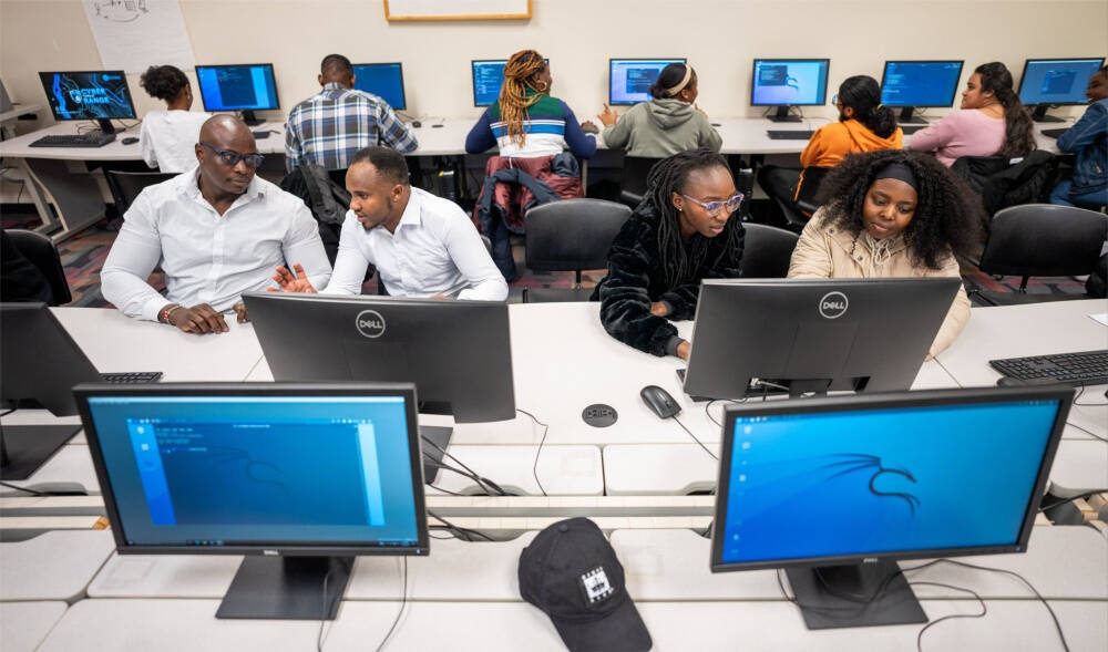 A classroom setting shows students working at desktop computers featuring the Kali Linux operating system. The students are arranged in pairs or groups, engaged in collaborative work, with a black baseball cap visible on one of the front tables.